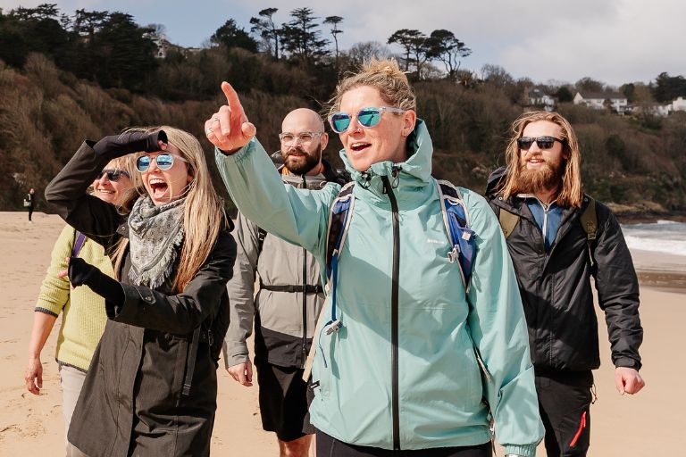 Group of five friends on a sandy beach, one person in a teal jacket points toward something in the distance while the others smile and wear sunglasses.