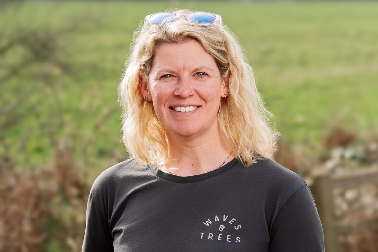Smiling blonde woman outdoors, wearing a black shirt with a 'WAVES & TREES' logo and sunglasses on her head.