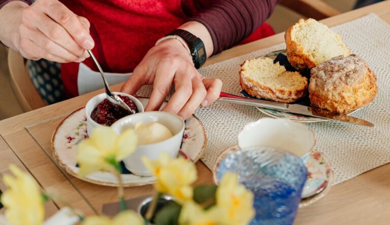 Person in a red sweater wearing a smartwatch slices a loaf cake at a tea-table, with jam, butter, and teacups nearby.