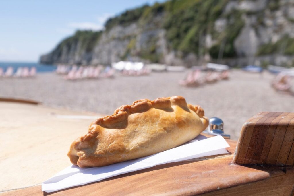 Golden baked pastry resting on a napkin atop a wooden table at a sunny beach, with lounge chairs and cliffs in the background.