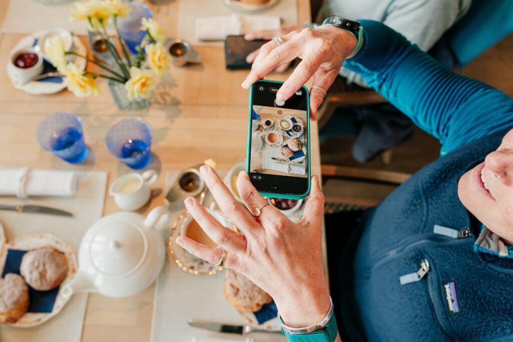 Hands hold a smartphone to photograph a breakfast table with teapot, pastries, cups and flowers.