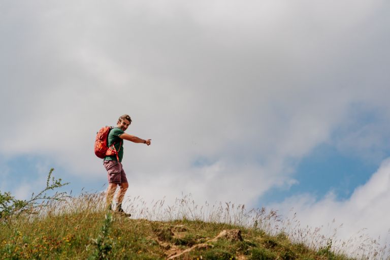 Hiker with an orange backpack stands on a grassy hill, pointing into the distance against a partly cloudy blue sky.