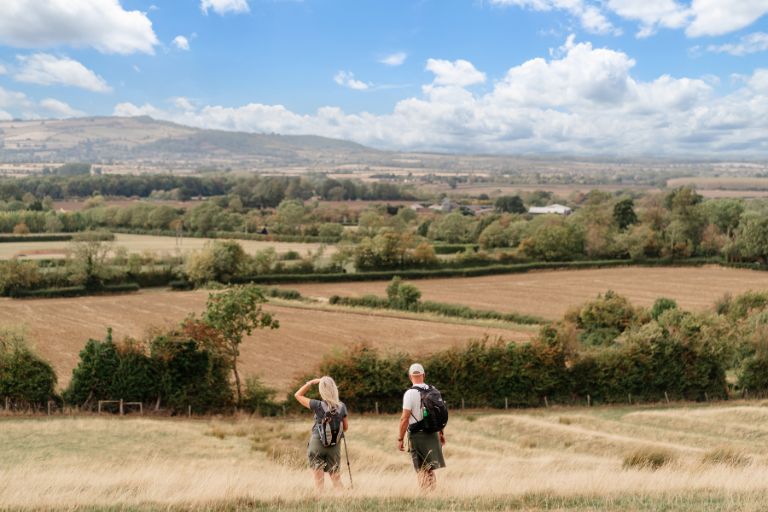 Two hikers with backpacks walk along a golden hillside overlooking a patchwork of fields under a blue sky with scattered clouds.