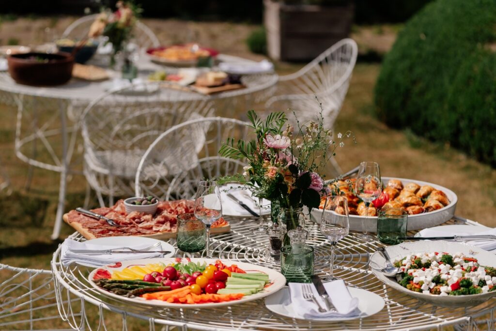 Outdoor metal table set for a meal with a floral centerpiece, wine glasses, and plates of vegetables, cheese, and bread in a garden setting.
