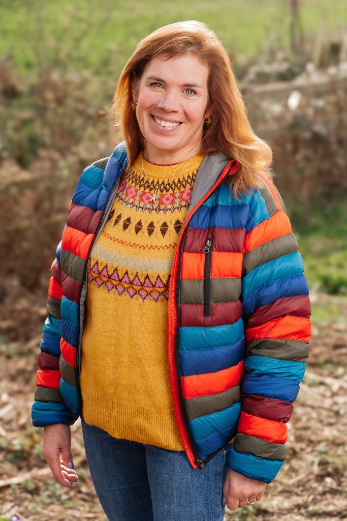 Smiling woman with light brown hair wearing a bright, striped puffer jacket outdoors in autumn light.