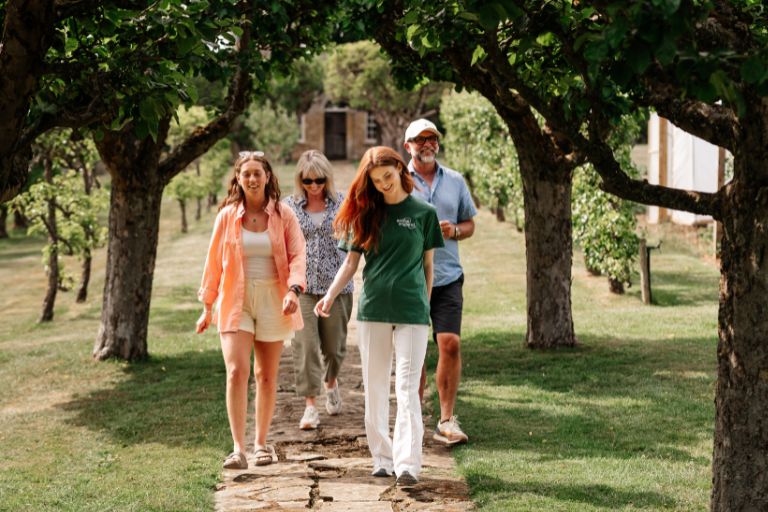 Group of five adults walking along a stone path through a park, shaded by trees and smiling.