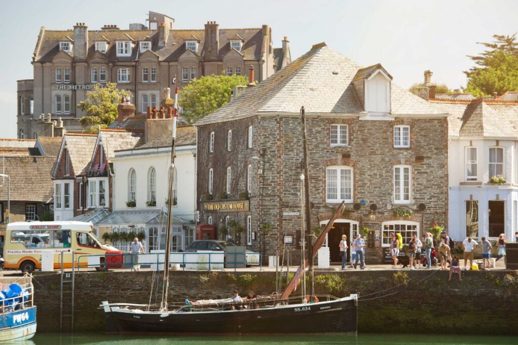 Quaint harbor town scene with stone buildings, a sailboat moored in the quay, and pedestrians along the waterfront.
