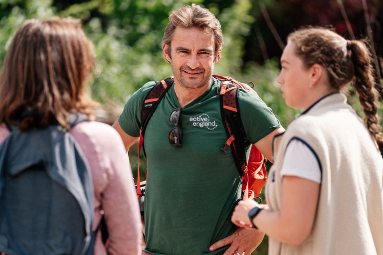 Man in green Active England shirt talks with two women hikers outdoors, backpacks on, sunny day.