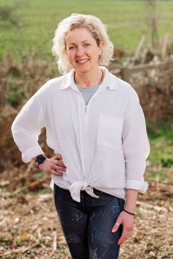 Smiling woman with blond curly hair in a white tied shirt and dark patterned jeans, standing outdoors in a vineyard setting.
