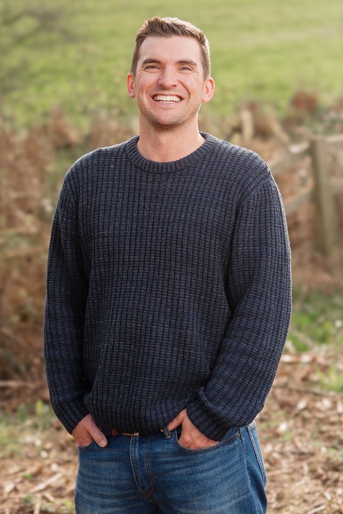 Smiling man in a dark knit sweater and jeans standing outdoors in a park-like setting, hands in pockets.